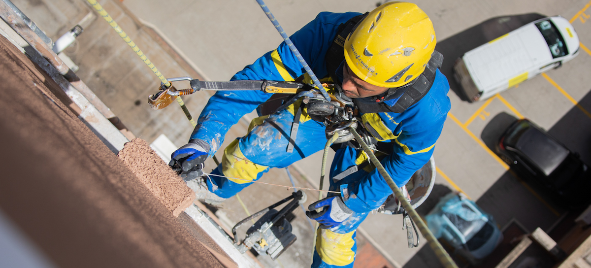 Ouvrier grimpant sur une corde vers un balcon pour réaliser des travaux de fermeture de terrasse sans autorisation.