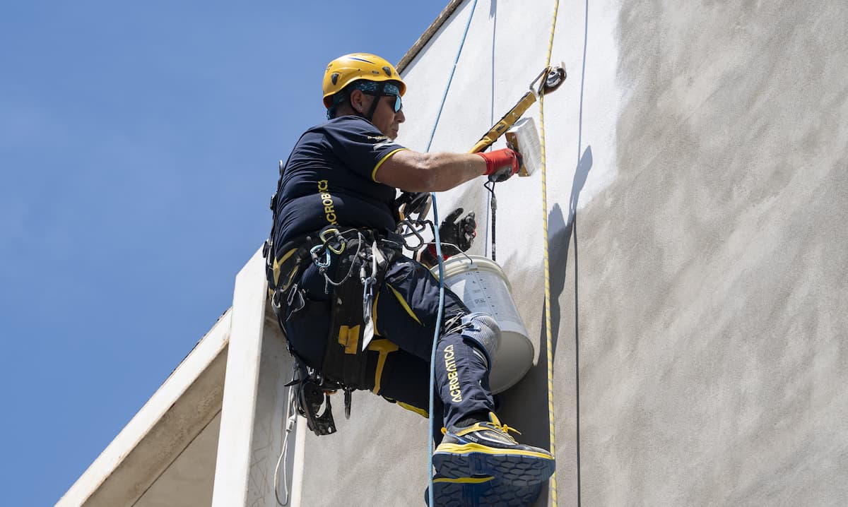 Technicien cordiste d'Acrobatica suspendu en hauteur à l'aide de cordes, en train de réparer et de préparer l'enduit d'un mur extérieur autour des fenêtres.